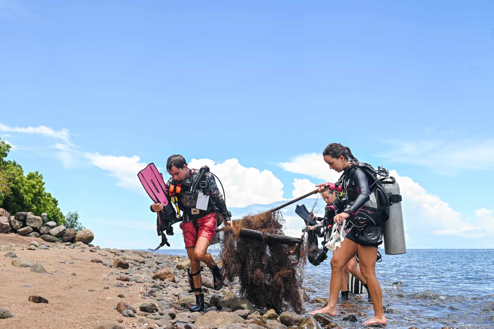 A picture of three divers walking on the beach, carrying a large discarded fishing net